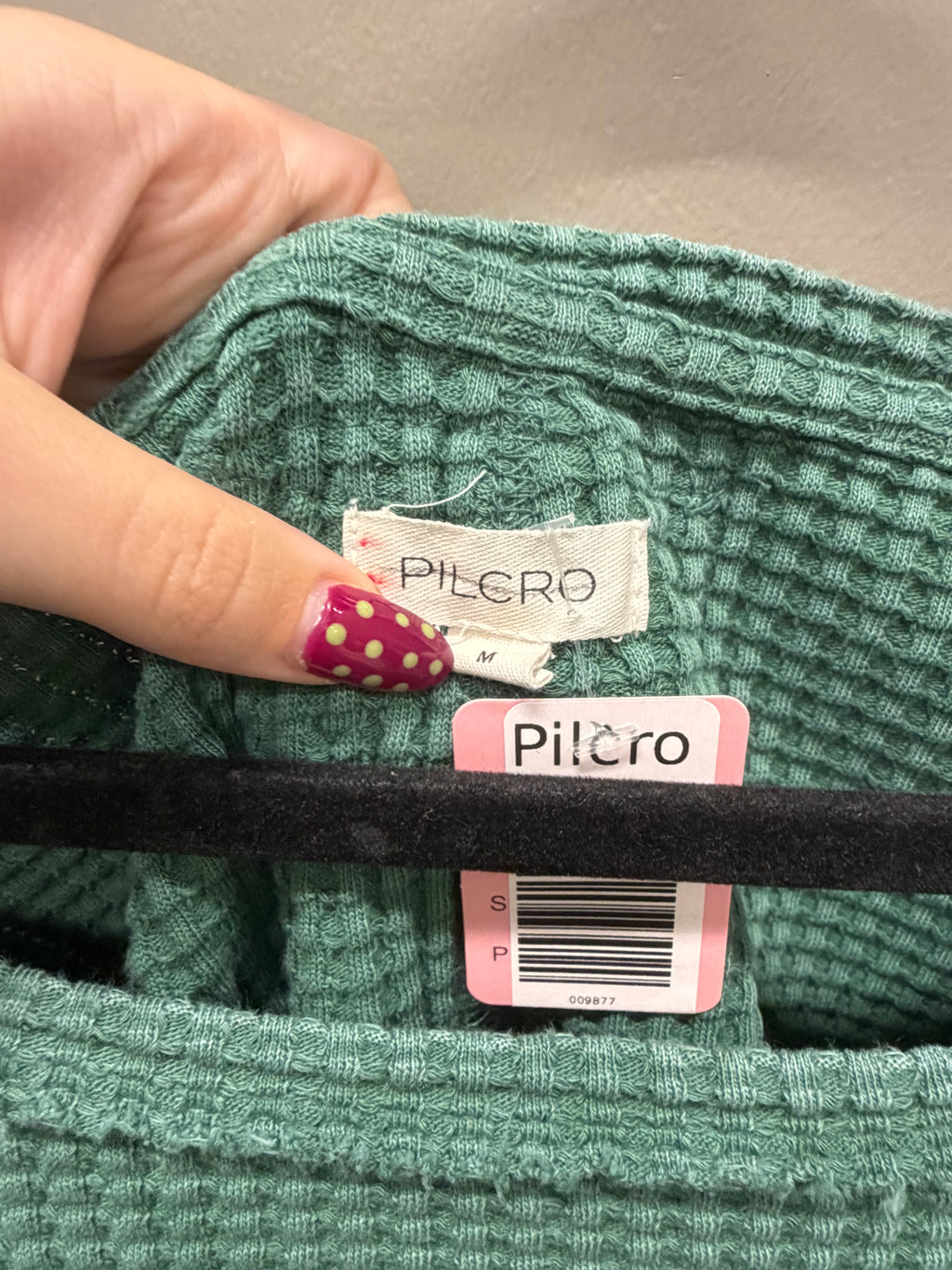 A close-up of a person's hand with pink and gold nail art holding a green ribbed knit garment open to show the Pilcro brand label and retail tag.