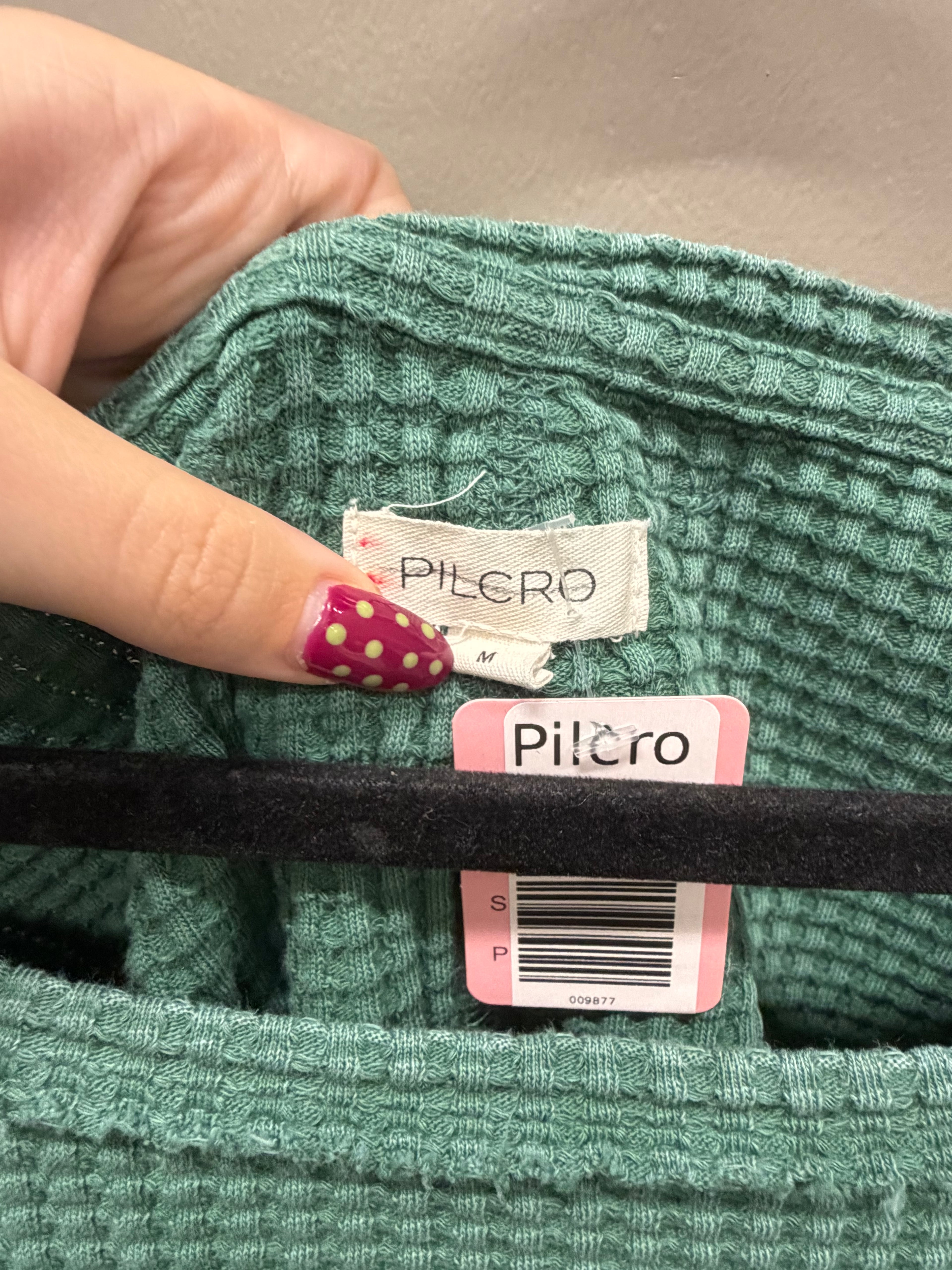 A close-up of a person's hand with pink and gold nail art holding a green ribbed knit garment open to show the Pilcro brand label and retail tag.