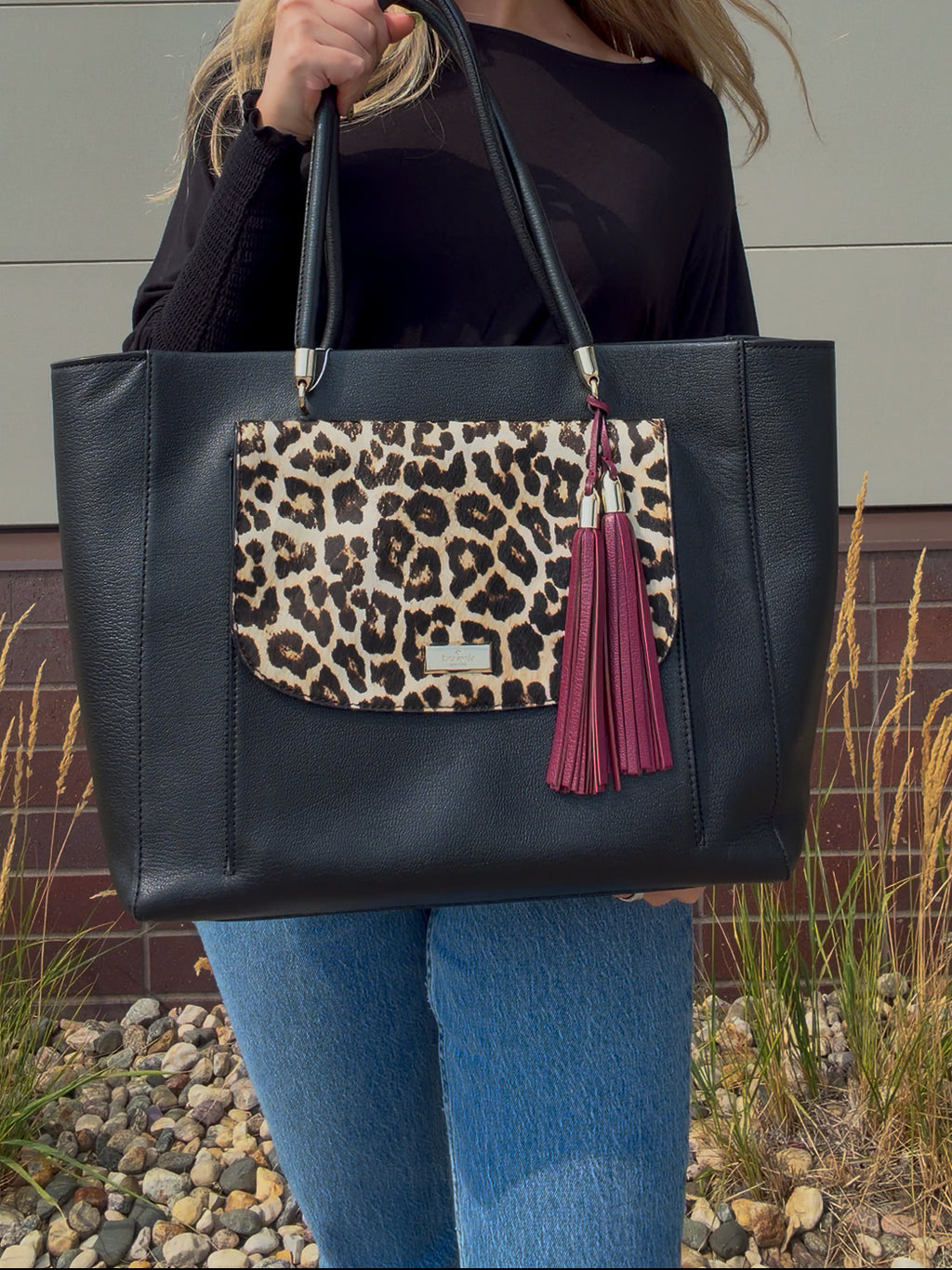 A person holding a large black faux-leather tote bag with a leopard-print front pocket and a burgundy tassel charm, shown outdoors against a neutral wall and gravel ground.