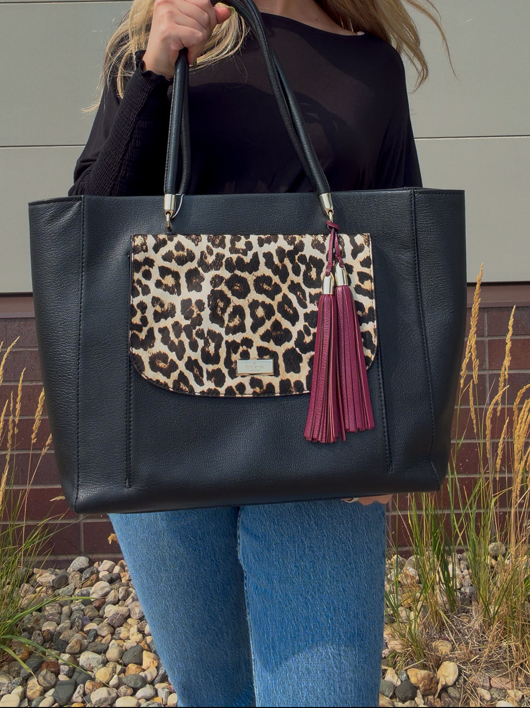A person holding a large black faux-leather tote bag with a leopard-print front pocket and a burgundy tassel charm, shown outdoors against a neutral wall and gravel ground.