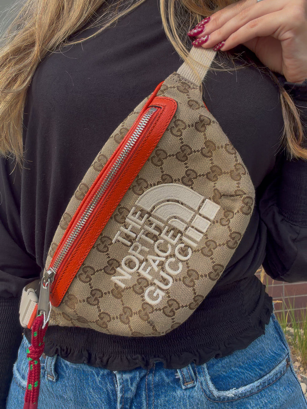 A close-up of a person wearing a beige GG Supreme canvas fanny pack with a bright orange-red leather zipper and a large white 'THE NORTH FACE' logo on the front.