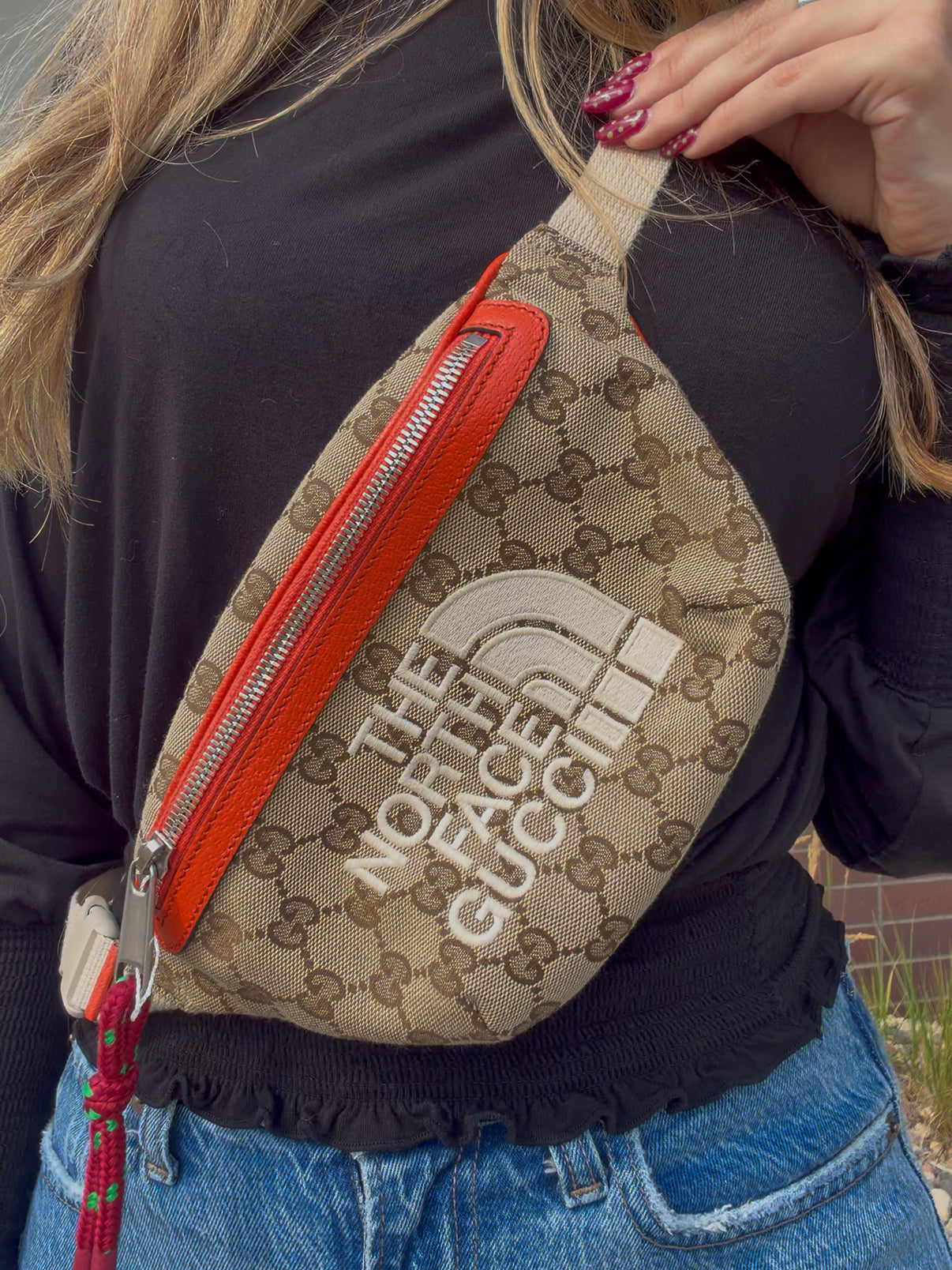 A close-up of a person wearing a beige GG Supreme canvas fanny pack with a bright orange-red leather zipper and a large white 'THE NORTH FACE' logo on the front.