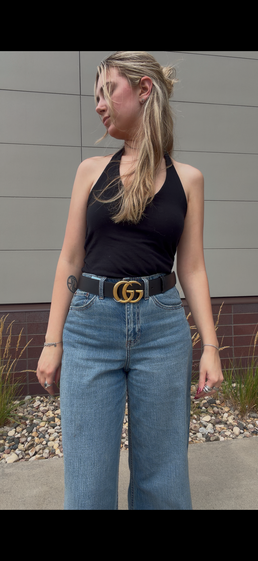A woman wearing a black halter top, wide-leg blue jeans, and a black belt with a gold interlocking  Gucci GG buckle, standing outdoors in front of a gray paneled wall.