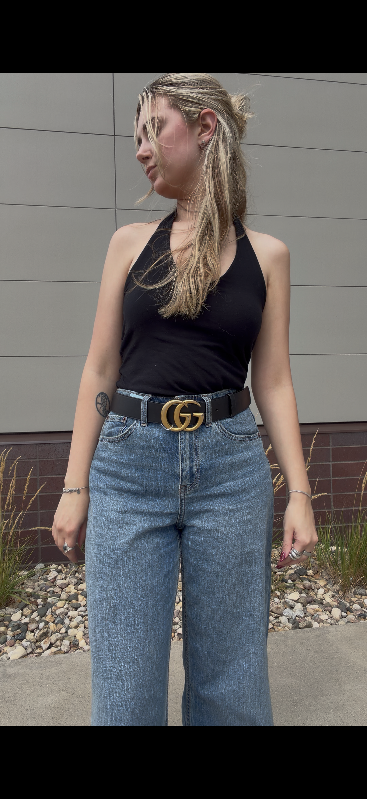 A woman wearing a black halter top, wide-leg blue jeans, and a black belt with a gold interlocking  Gucci GG buckle, standing outdoors in front of a gray paneled wall.