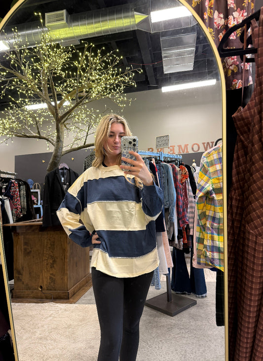 A woman takes a mirror selfie in a clothing boutique wearing a loose, oversized long-sleeve striped top with navy blue and cream horizontal bands, paired with black leggings.