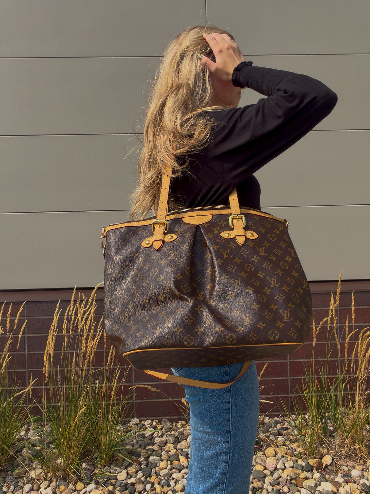A woman shown from behind carrying a large Louis Vuitton monogram Neverfull tote bag over her shoulder, standing outdoors in front of a gray paneled wall.
