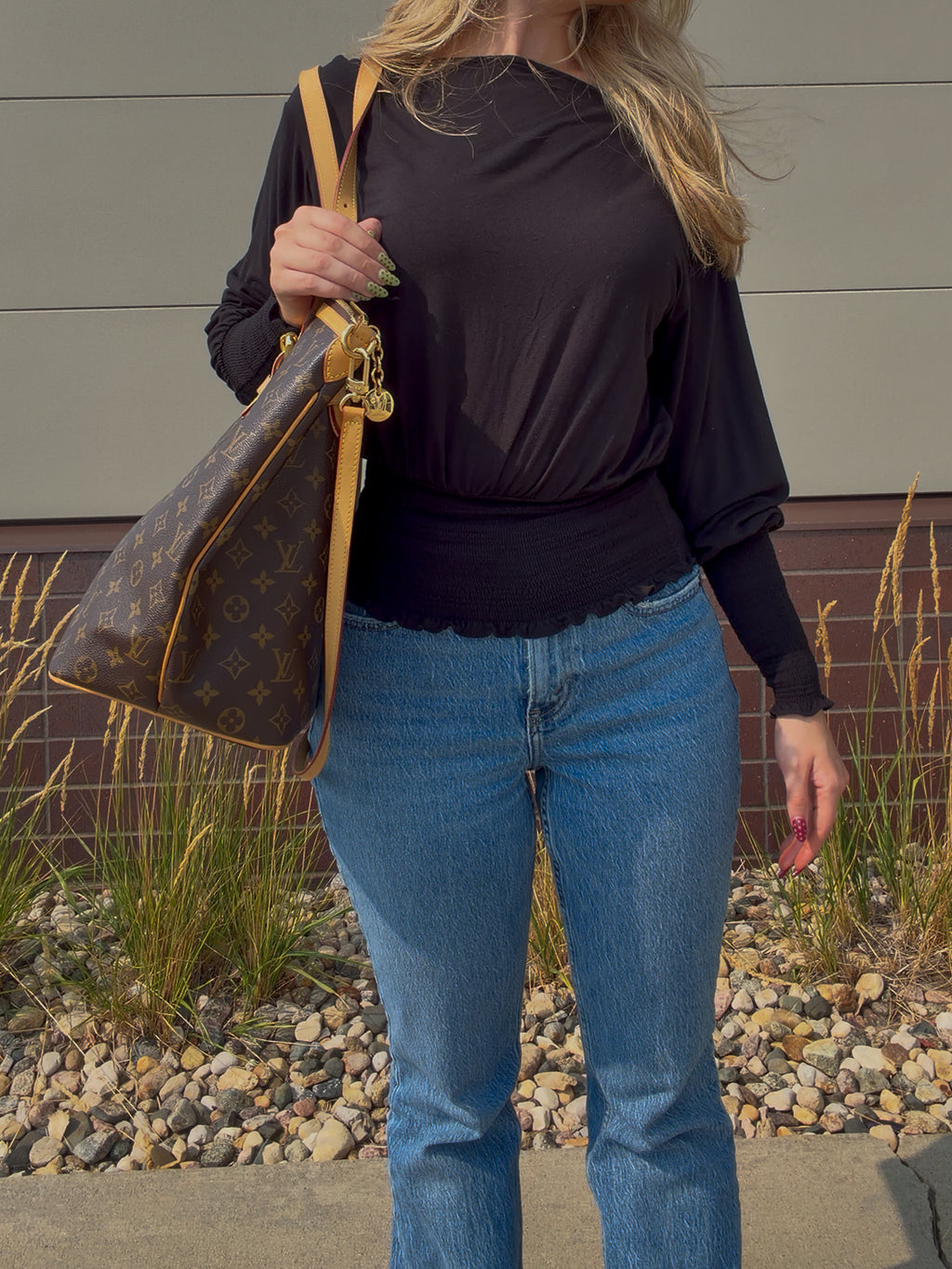 A woman wearing a black long-sleeve top and blue jeans carries a brown Louis Vuitton monogram shoulder bag with tan leather trim in an outdoor setting.