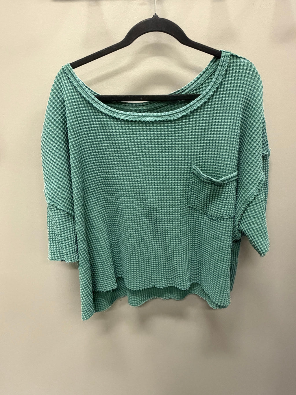 A green and white micro-check patterned waffle-knit top with a scoop neckline, 3/4 sleeves, and a chest pocket, hanging on a black hanger against a plain beige wall.
