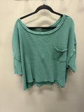 A green and white micro-check patterned waffle-knit top with a scoop neckline, 3/4 sleeves, and a chest pocket, hanging on a black hanger against a plain beige wall.