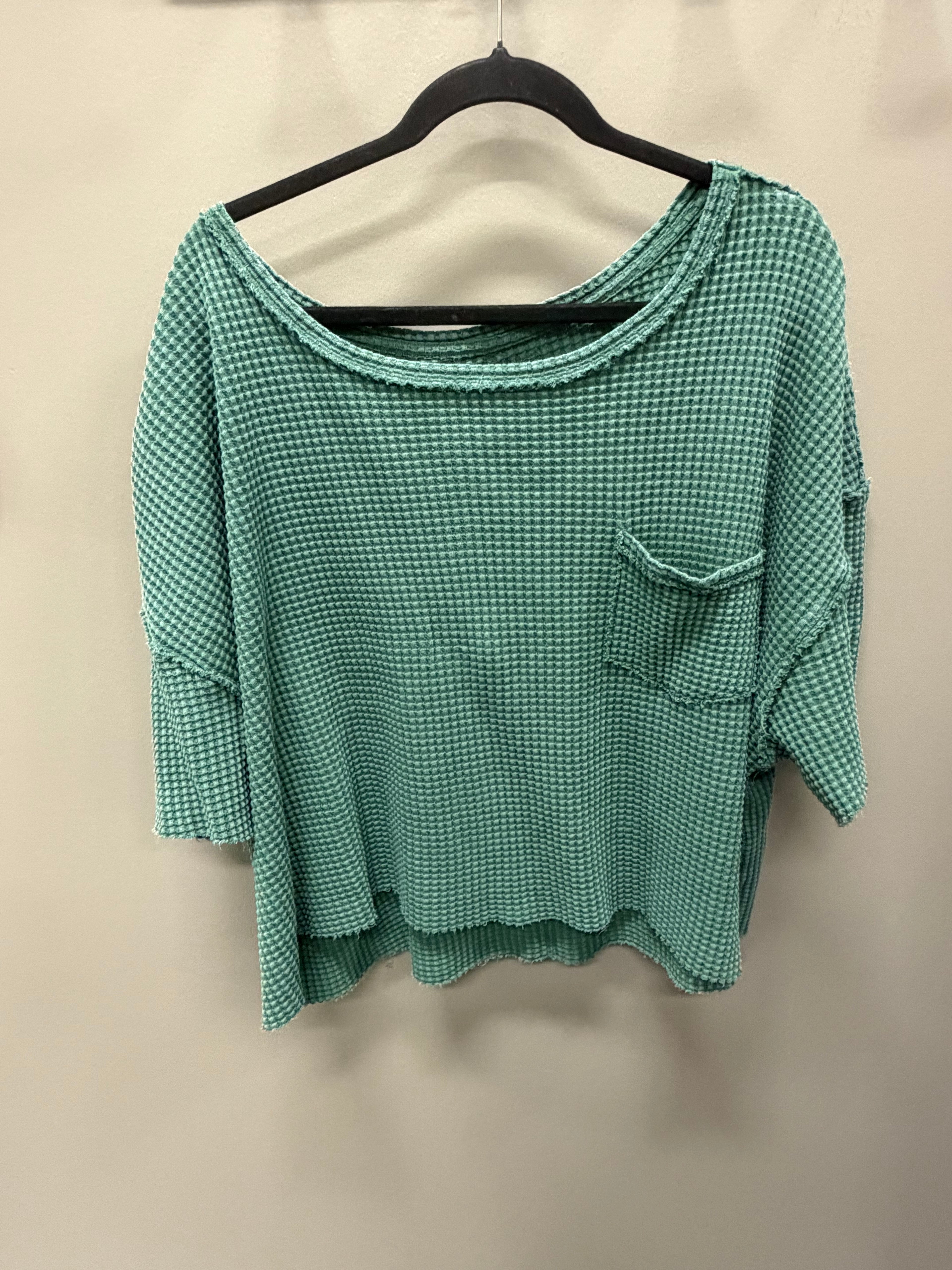 A green and white micro-check patterned waffle-knit top with a scoop neckline, 3/4 sleeves, and a chest pocket, hanging on a black hanger against a plain beige wall.