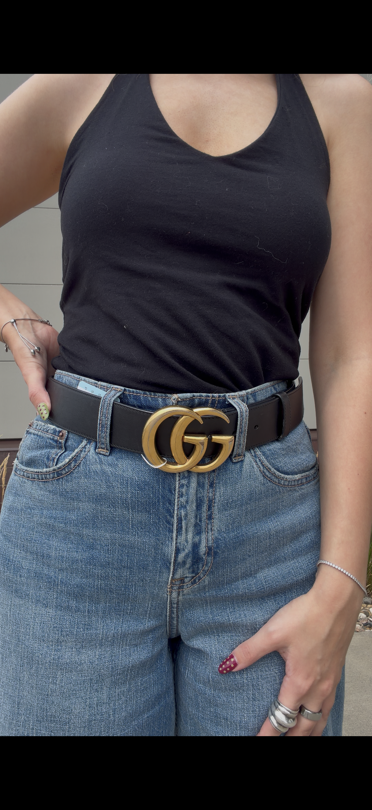 A close-up of a person wearing a black Gucci belt with a prominent gold interlocking GG logo buckle, styled with a black tank top and blue jeans.