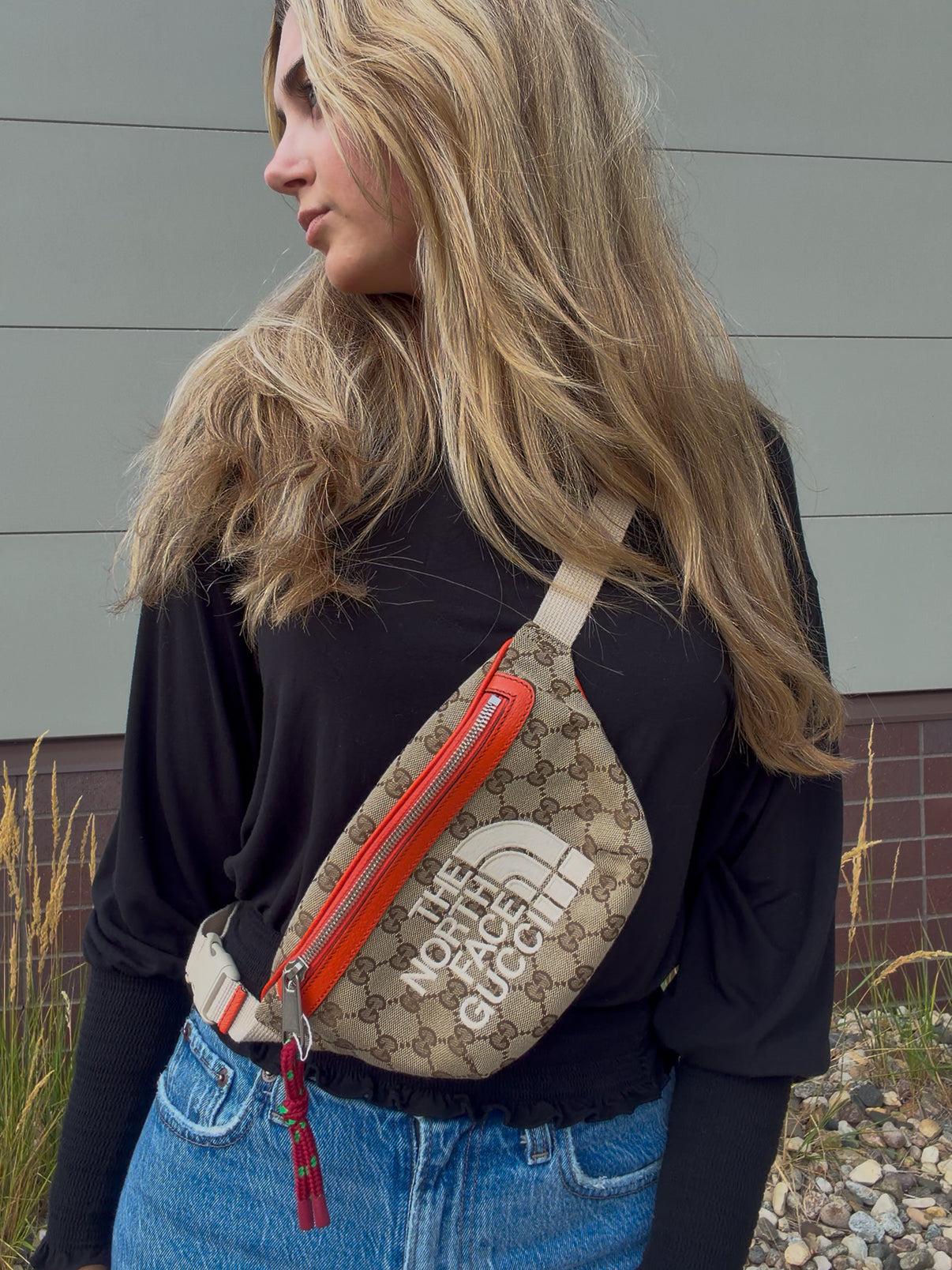 A woman wearing a beige and brown GG Supreme crossbody bag with a prominent orange zipper and a white 'THE NORTH FACE' logo, styled with a black long-sleeve shirt and blue jeans.
