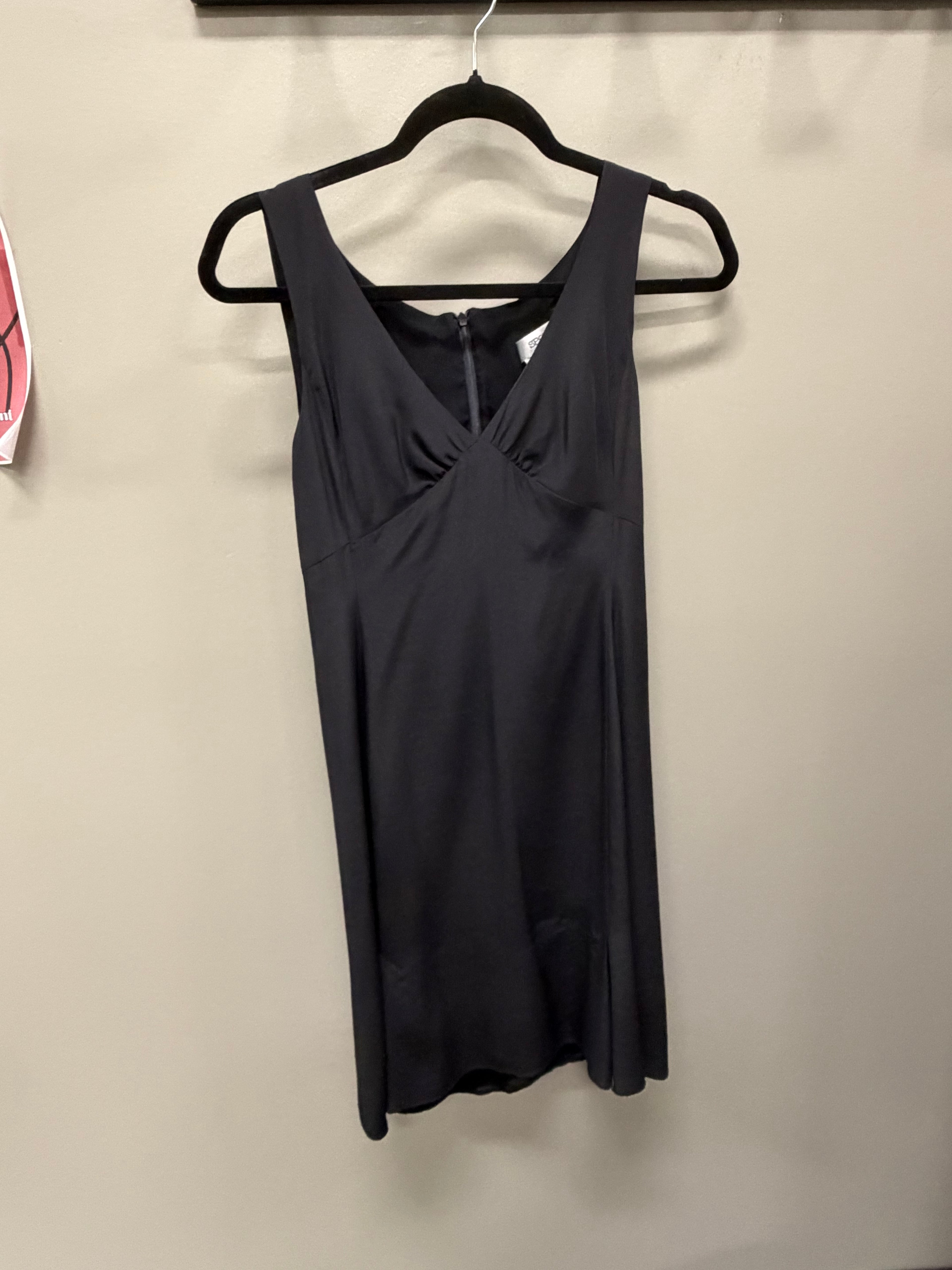 A sleeveless black dress with a V-neckline and gathered front detail, hanging on a black hanger against a plain beige wall.