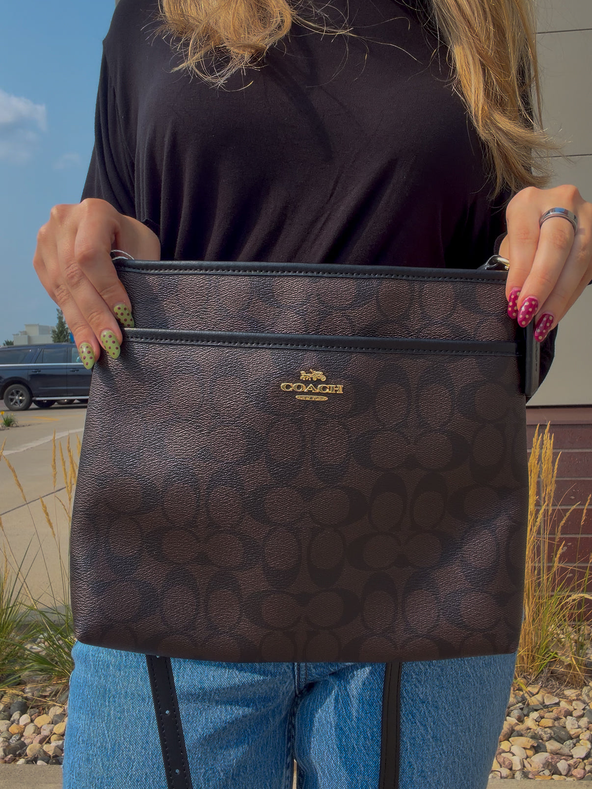 A person holding a brown Coach crossbody bag with the signature monogram pattern and gold logo. The person is wearing a black top and blue jeans, with an outdoor background visible.