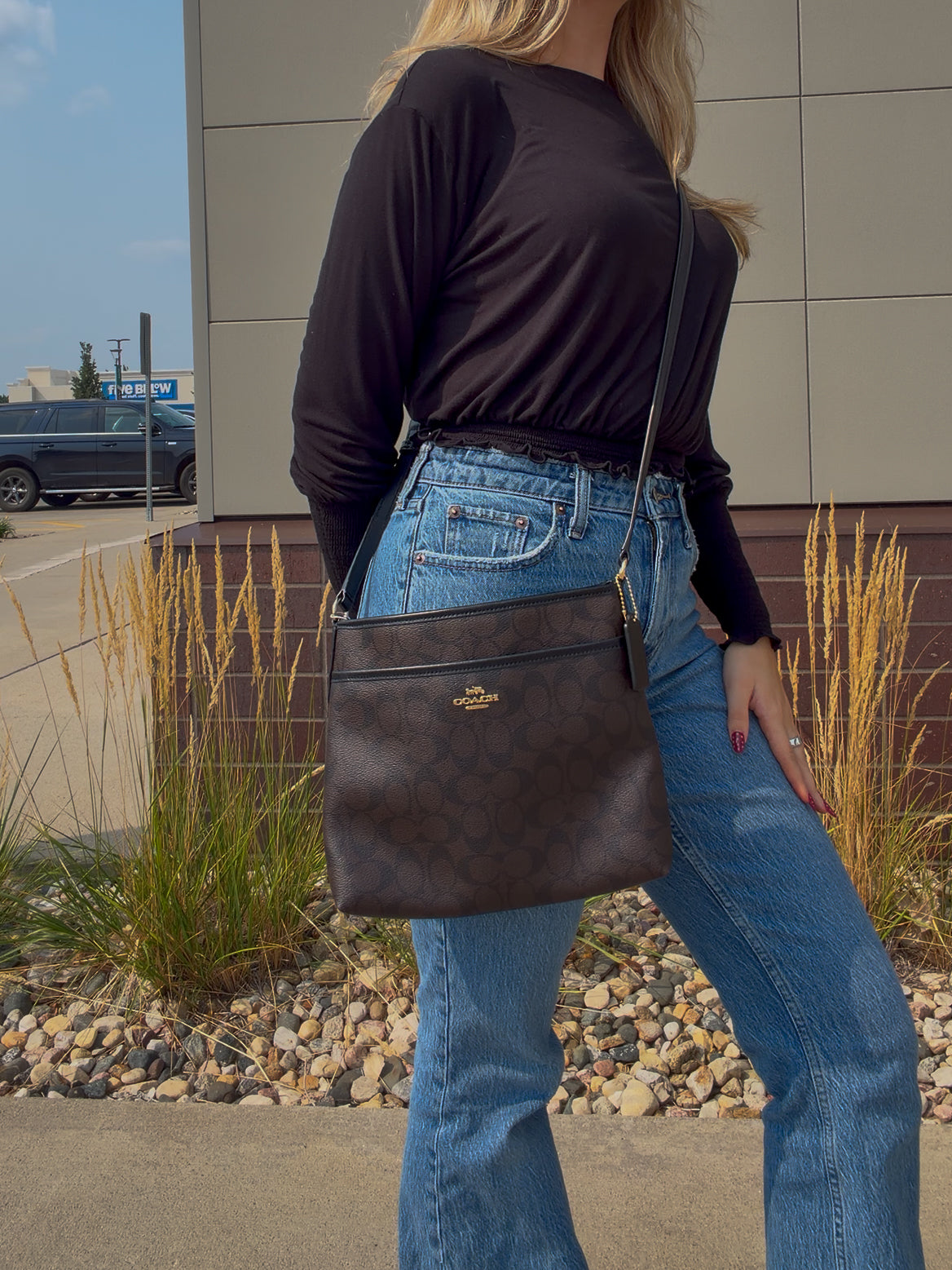 A woman wearing a black long-sleeve top and blue jeans carries a dark brown monogram crossbody bag with a gold-tone logo on a city street.