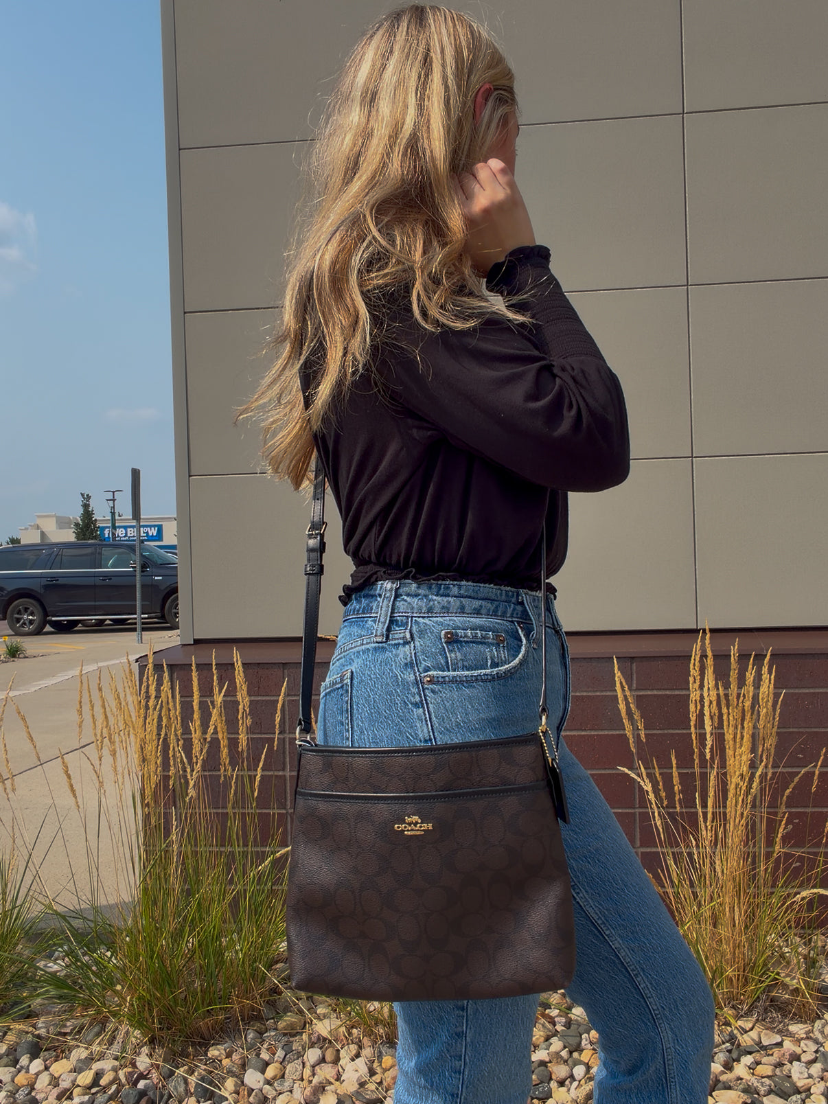 A woman wearing a black long-sleeve top and blue jeans is shown from the back, carrying a dark brown monogram crossbody bag with a gold logo on a city street next to a modern building.