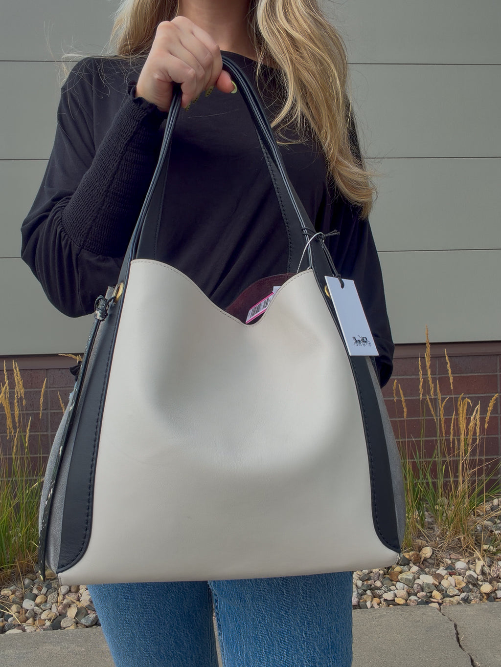 A person holding a large hobo-style shoulder bag outdoors. The bag is off-white/cream with black side panels and black handles.