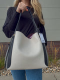 A person holding a large hobo-style shoulder bag outdoors. The bag is off-white/cream with black side panels and black handles.