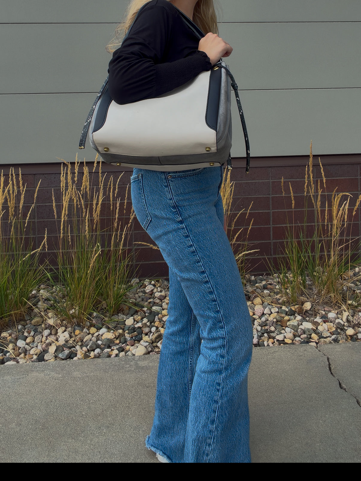 A person wearing blue flared jeans and a black long-sleeve top carries a large white and gray shoulder bag with dark trim, standing outdoors in front of a gray wall and gravel planter bed.