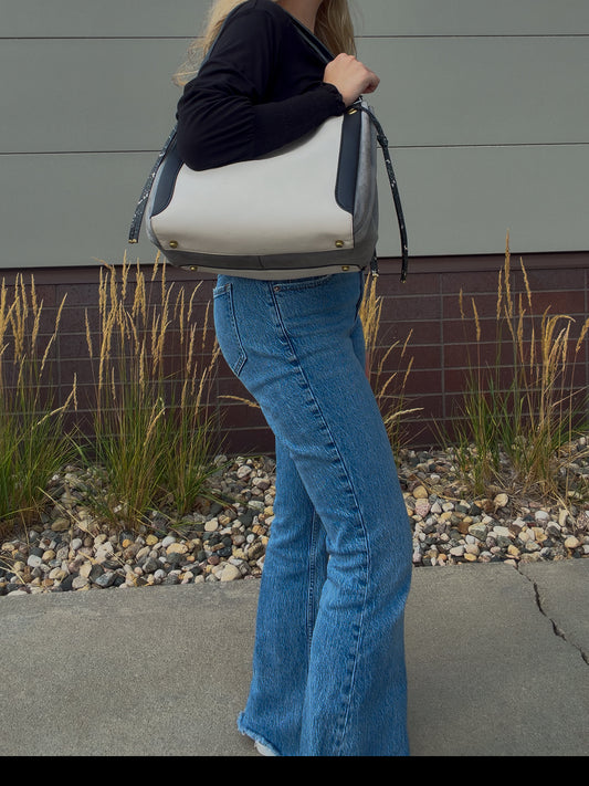 A person wearing blue flared jeans and a black long-sleeve top carries a large white and gray shoulder bag with dark trim, standing outdoors in front of a gray wall and gravel planter bed.