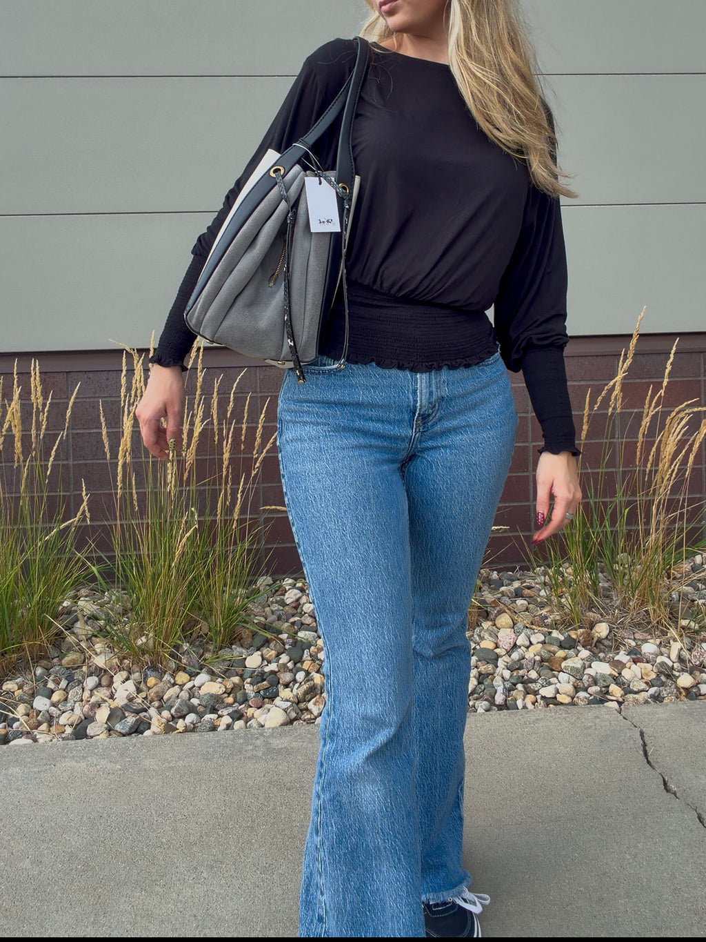 A woman wearing a black long-sleeve top and light blue flare jeans, carrying a gray shoulder bag outdoors against a gray wall and gravel background.