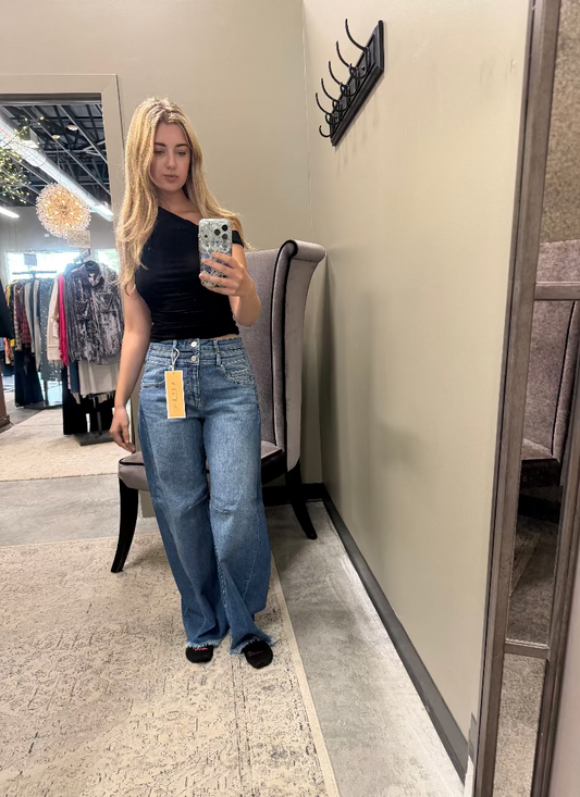 A woman takes a mirror selfie in a boutique wearing a black off-the-shoulder top and wide-leg blue jeans with a yellow price tag attached. The boutique interior features a gray upholstered chair, a beige wall, and clothing racks in the background.