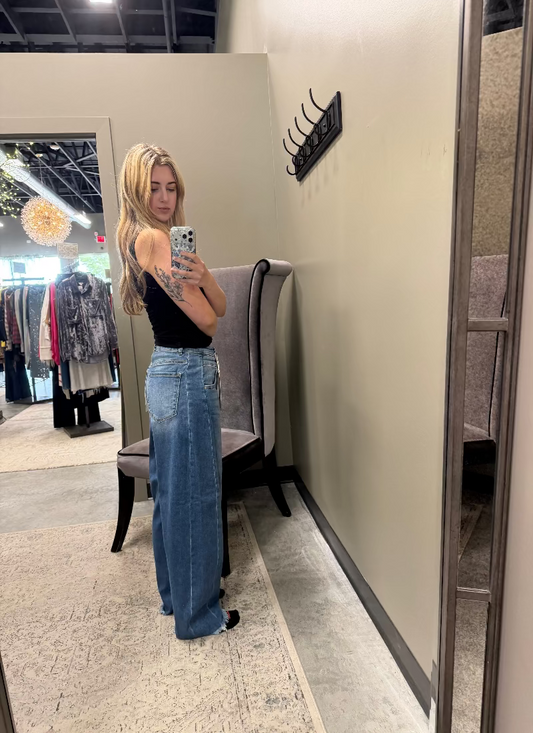 A woman taking a mirror selfie in a boutique, wearing a black sleeveless top and high-waisted, wide-leg blue jeans.