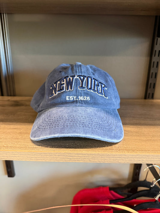 A blue washed cotton baseball cap with 'NEW YORK EST. 1625' embroidered on the front, resting on a wooden shelf.