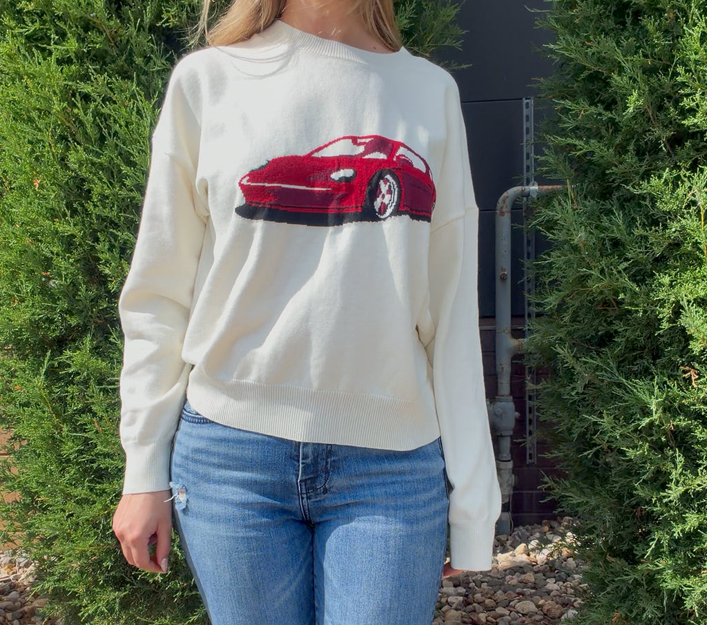 Person wearing a white sweater with a red car graphic outdoors.