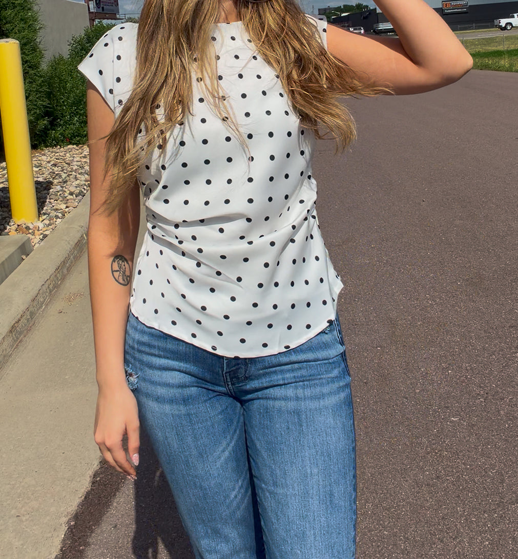 Person wearing a white polka dot top and blue jeans on a street.