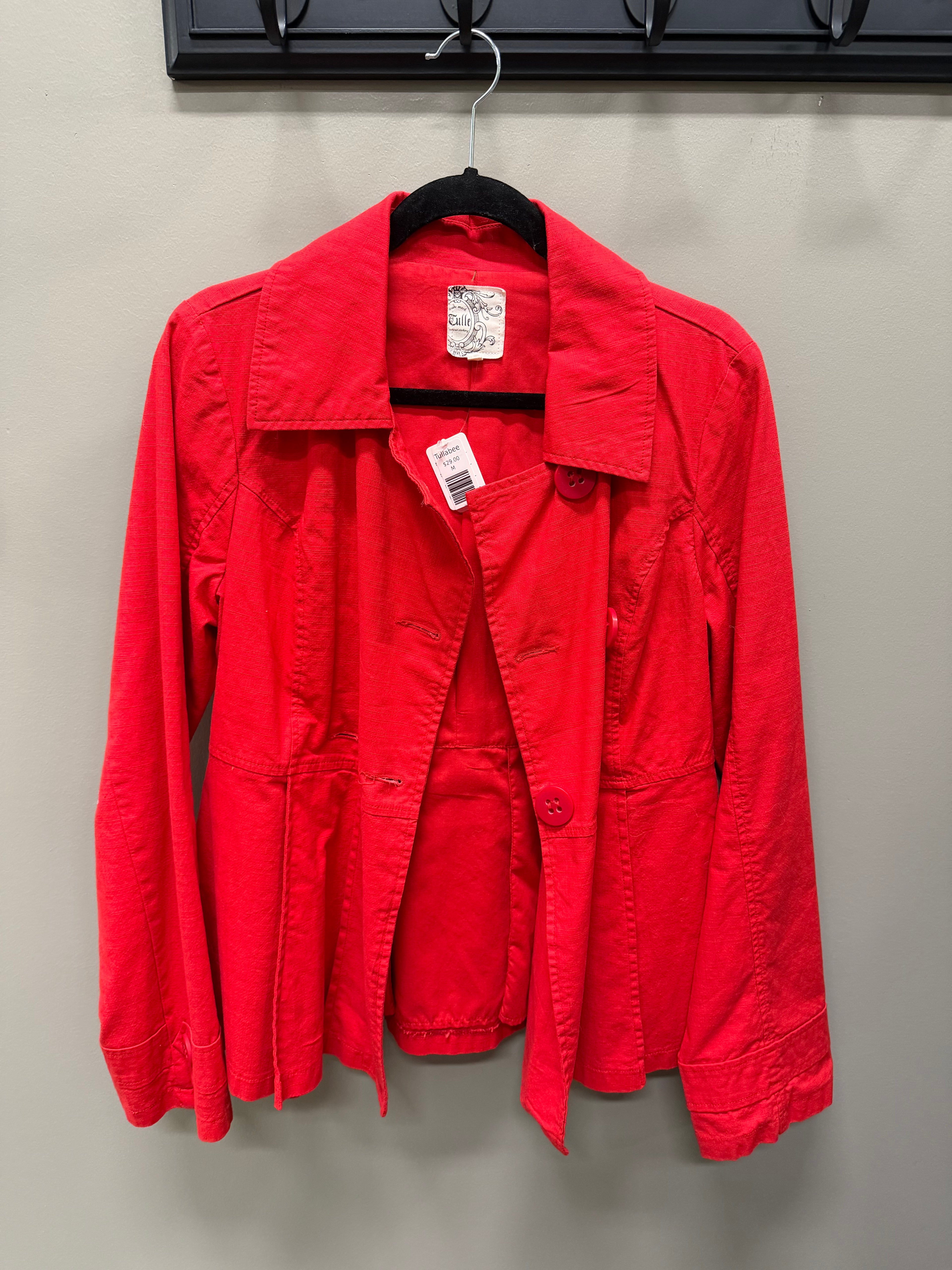 A bright red utility jacket hanging on a black hanger against a light gray wall. The jacket has a pointed collar, long sleeves, and front button closure, with a visible white retail price tag and a brand label inside the collar.