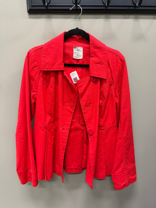 A bright red utility jacket hanging on a black hanger against a light gray wall. The jacket has a pointed collar, long sleeves, and front button closure, with a visible white retail price tag and a brand label inside the collar.