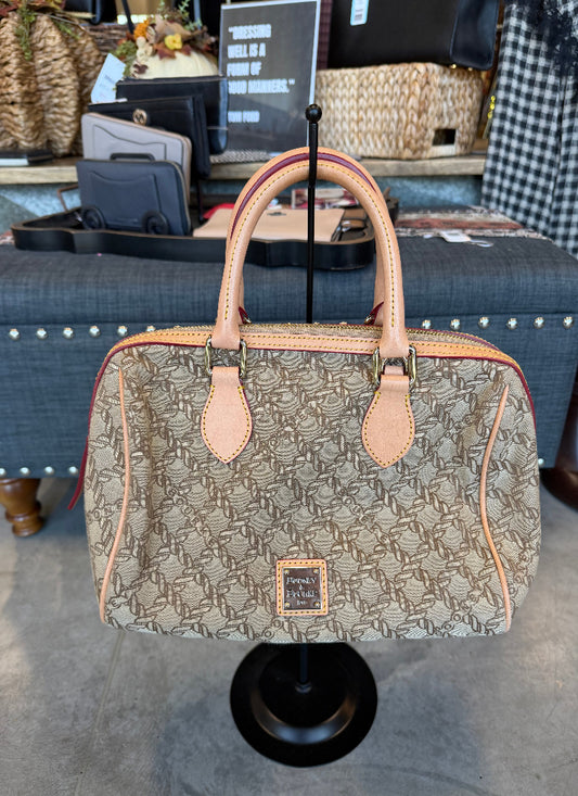 A beige and brown monogram handbag with light tan leather handles and trim, displayed on a black stand in a retail store setting.