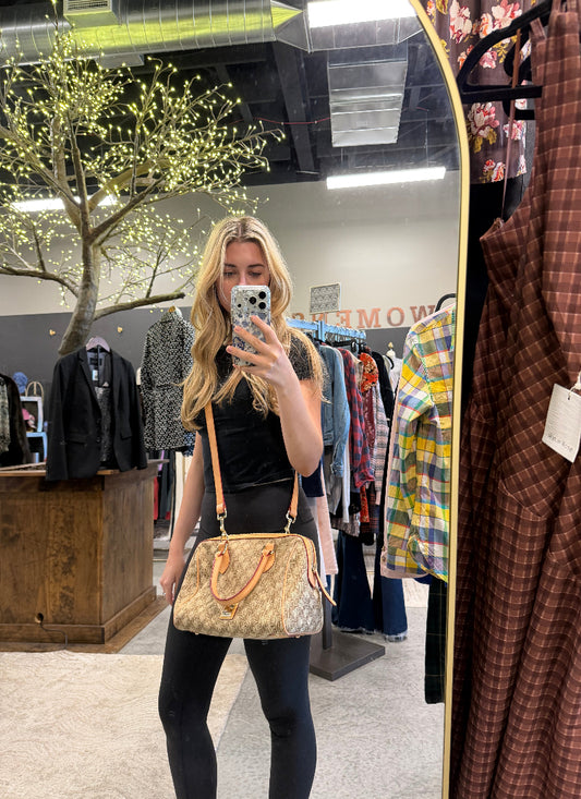 A woman takes a mirror selfie in a clothing store while wearing a beige and brown monogram crossbody handbag with tan leather straps. The background shows racks of clothes, a decorative tree with lights, and store fixtures.