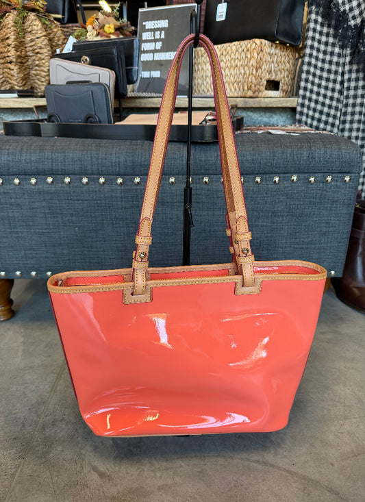 A glossy coral orange patent leather tote bag with tan leather handles and trim, displayed on a black stand in a retail store setting.