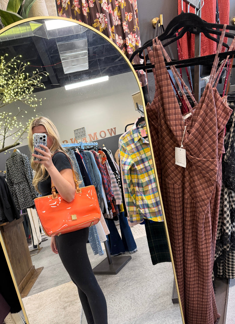 A woman takes a mirror selfie in a clothing boutique, holding a bright orange patent leather handbag. The boutique features a gold-framed mirror, racks of various patterned clothing, and a gray wall in the background.