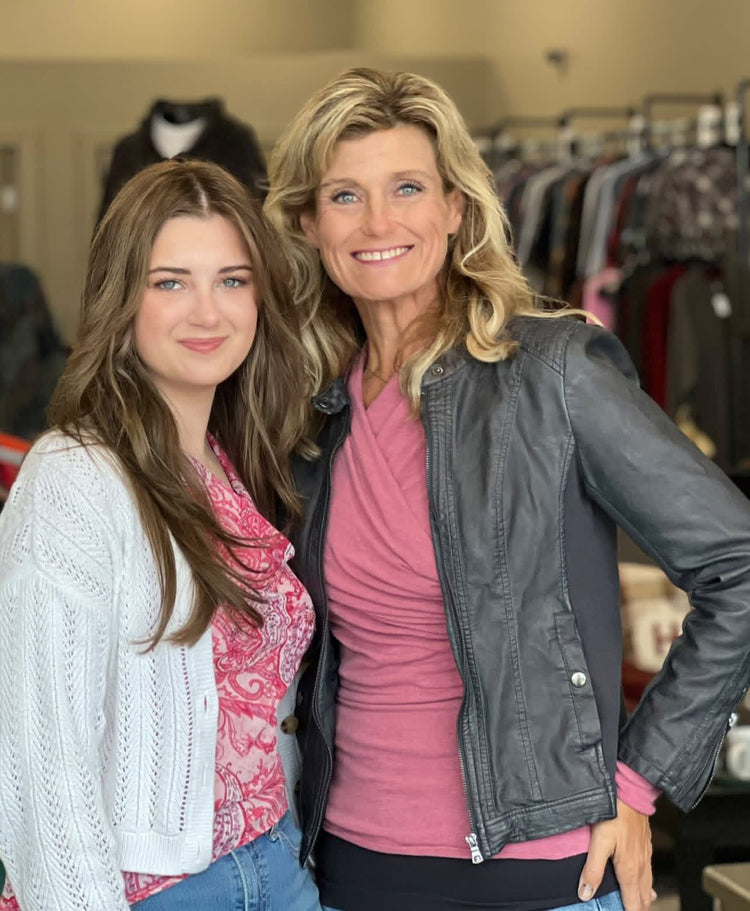 Two women posing together in a clothing store. The woman on the right wears a black faux-leather jacket over a pink top, and the woman on the left wears a white knit cardigan over a pink patterned blouse.