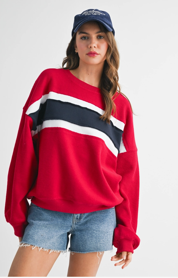 A woman wearing a red crewneck sweatshirt with diagonal navy and white stripes across the chest, paired with denim shorts and a navy baseball cap, against a plain light gray background.