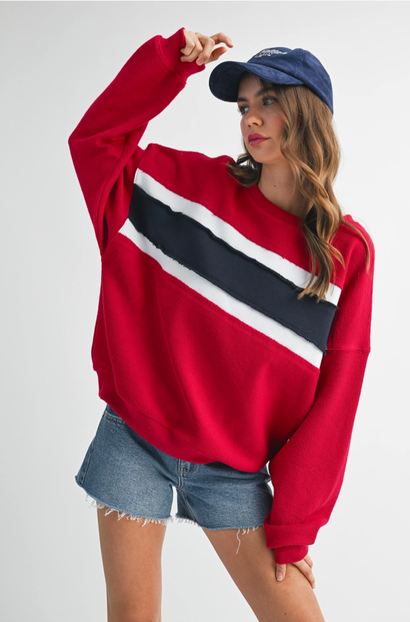 A woman modeling a red crewneck sweatshirt with a bold diagonal stripe pattern in navy and white across the chest, paired with denim shorts and a navy baseball cap against a light gray studio background.
