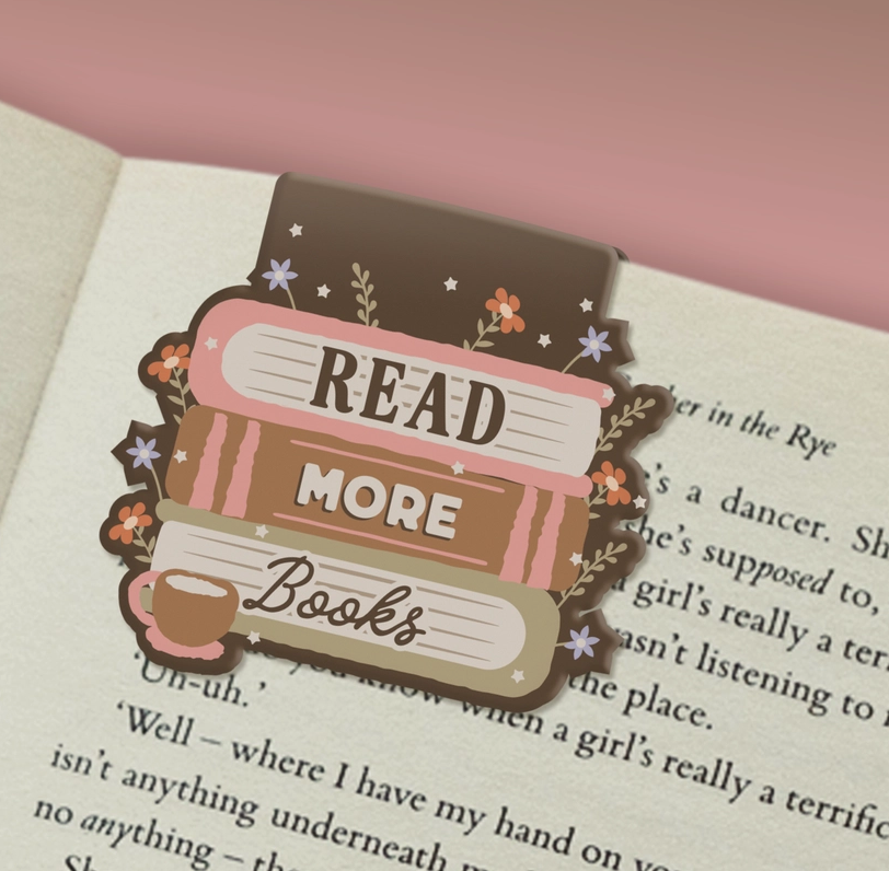 A decorative book-shaped bookmark with the phrase “READ MORE Books” sits on an open book. The bookmark features stacked illustrated books in pink, brown, cream, and beige, with small floral and star accents, plus a small coffee cup illustration.