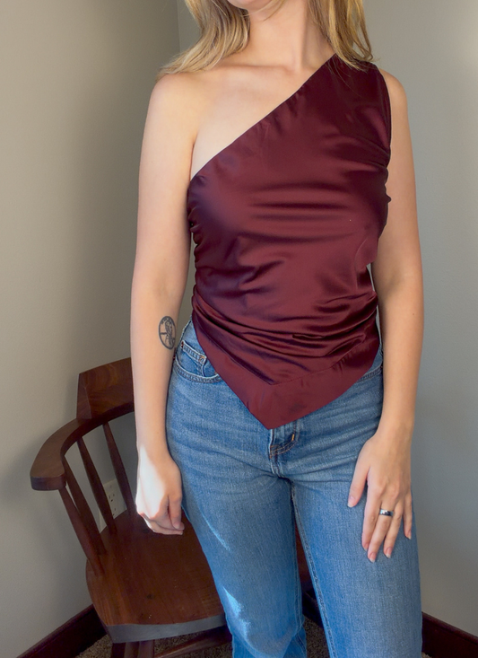 A woman wearing a satin one-shoulder top in deep burgundy with blue jeans, standing indoors next to a wooden chair.