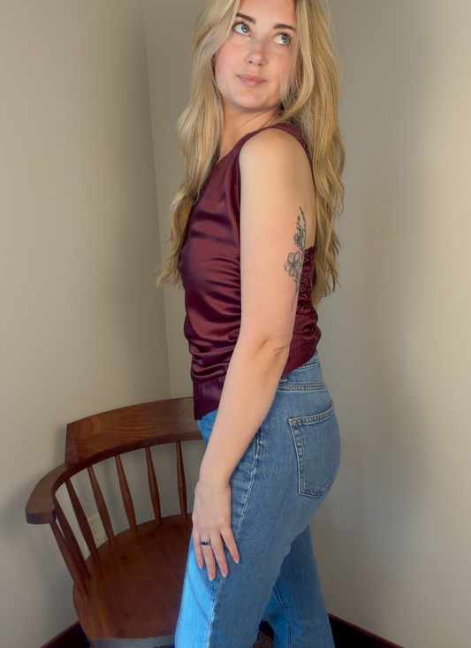 A woman with blonde hair stands in a corner wearing a sleeveless satin-like burgundy top and blue jeans, with a wooden chair visible behind her.