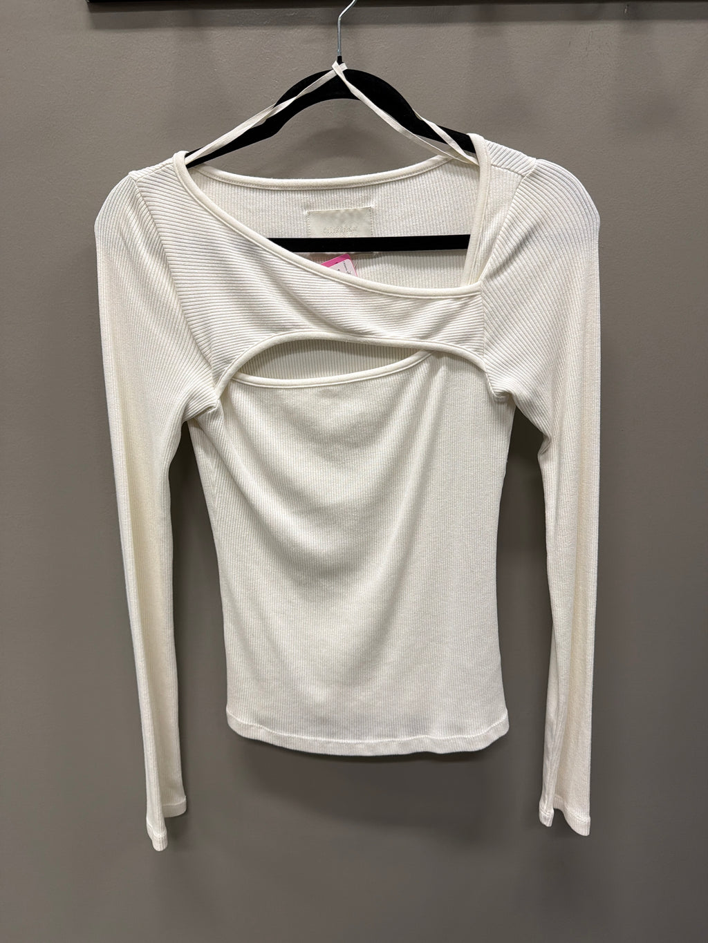 A white long-sleeve ribbed knit top with a unique cutout design on the chest, hanging on a black hanger against a dark gray wall.